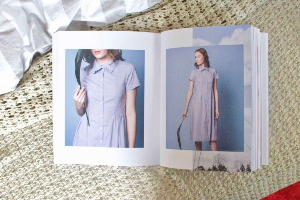An open book showcases spring sewing ideas. Both pages feature a woman with long hair wearing a light gray dress, set against a blue sky background. The book rests on a textured, cream-colored surface.
