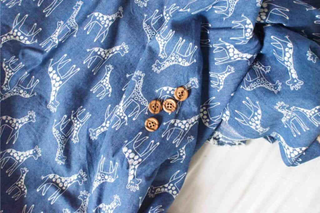Blue fabric with a white giraffe print pattern, featuring several wooden buttons scattered on top. The fabric and buttons are laid out on a light-colored surface.