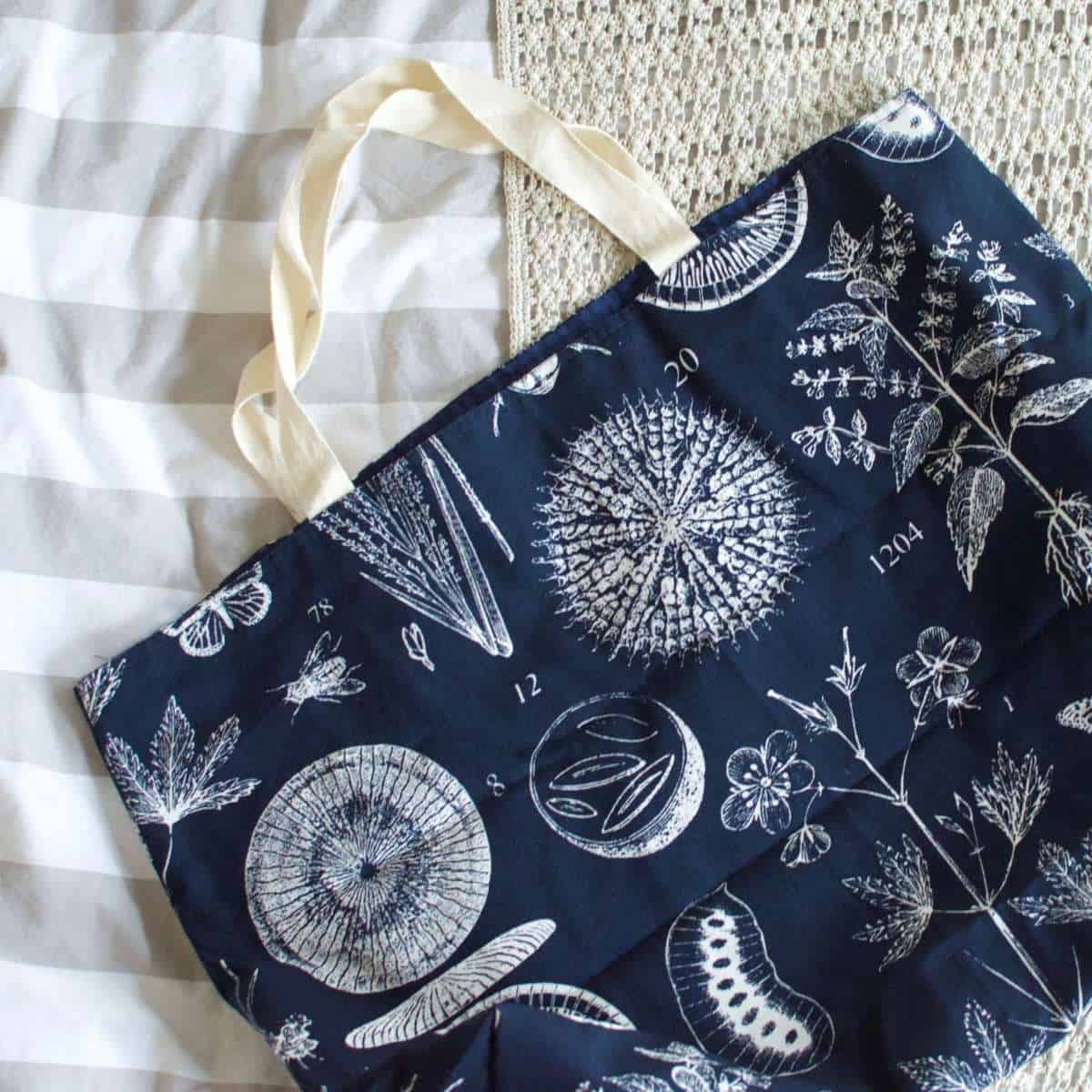 A blue tote bag adorned with white illustrations of plants and scientific diagrams rests on a textured, striped surface. Its cream-colored handles add charm to this creation, perfect for anyone crafting their spring sewing plans.