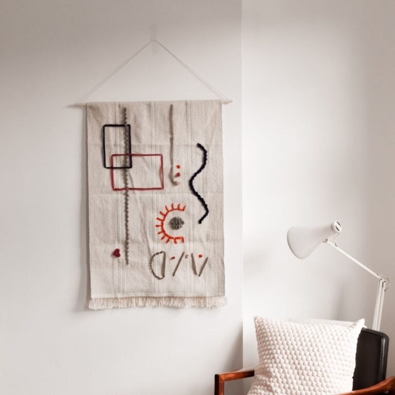 Make a tapestry wall hanging with abstract geometric shapes in black, orange, and red on a cream background. Its displayed above a chair with a white pillow. A white lamp is nearby, all set against a light grey wall.