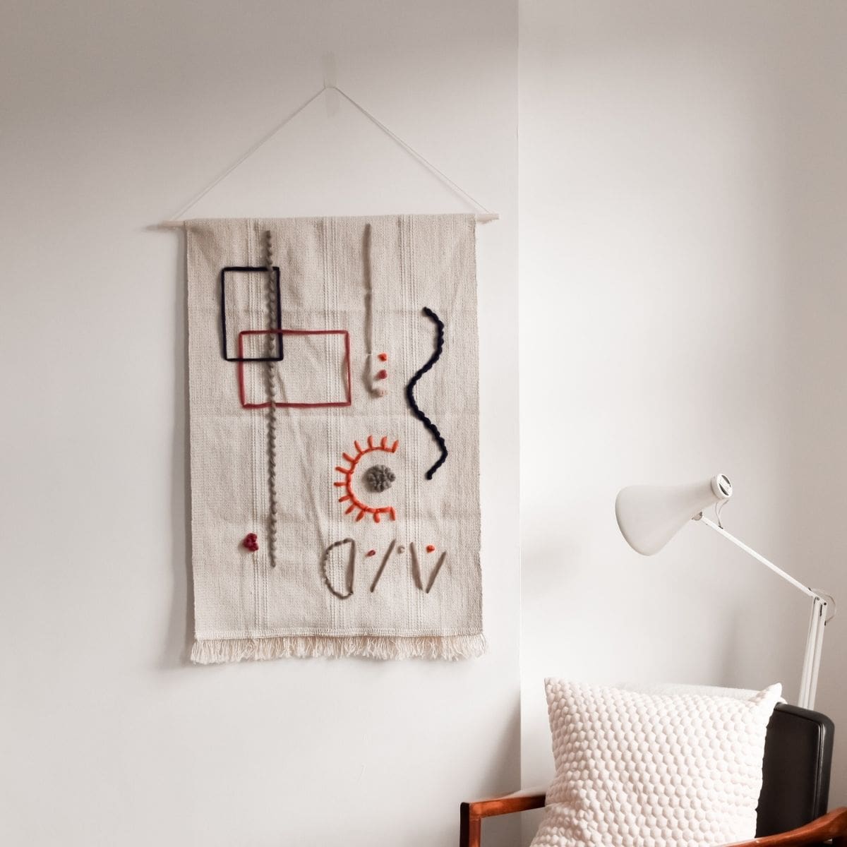 Make a tapestry wall hanging with abstract geometric shapes in black, orange, and red on a cream background. Its displayed above a chair with a white pillow. A white lamp is nearby, all set against a light grey wall.