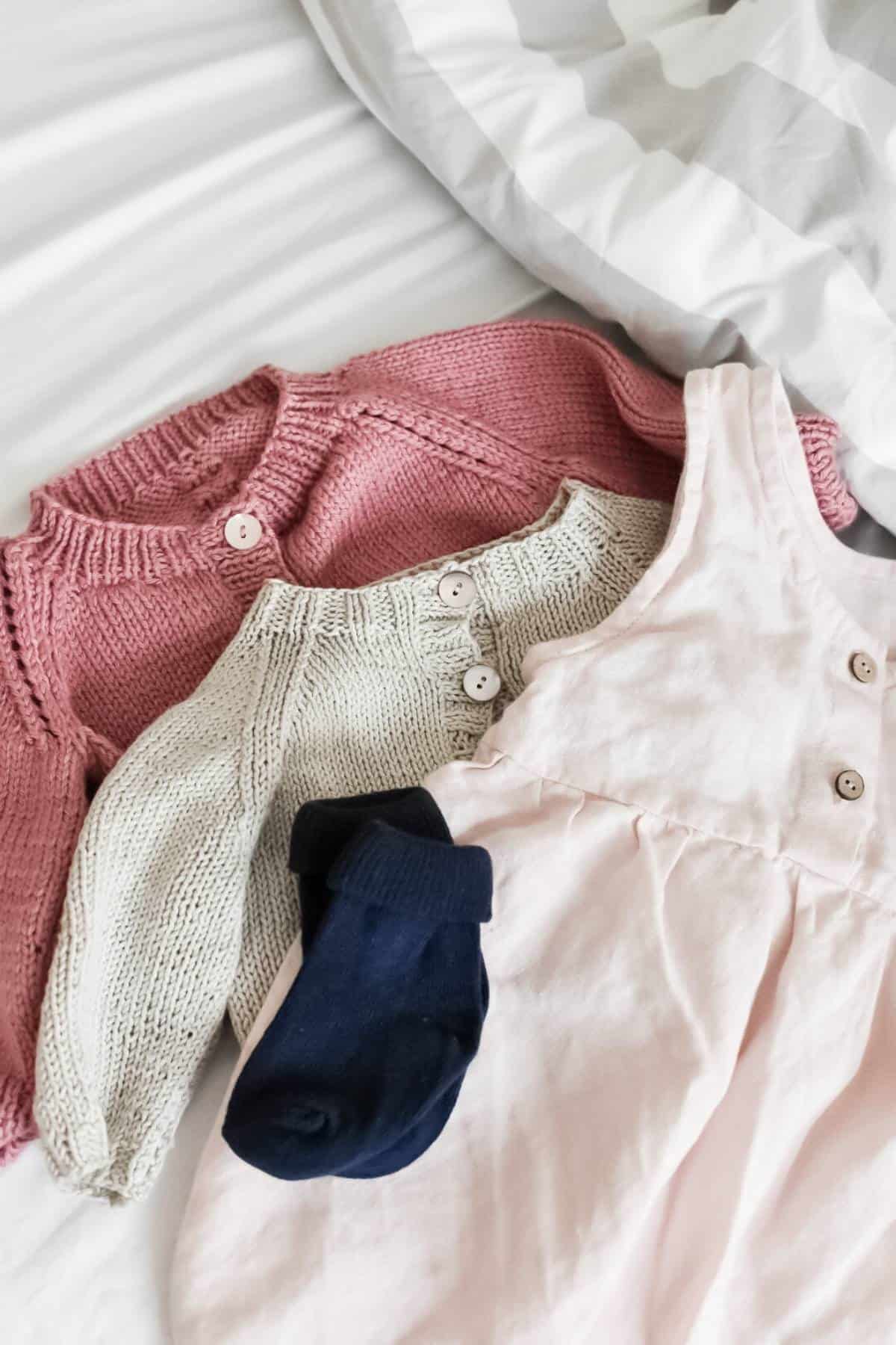 Childrens clothing laid out on a bed: a pink knitted sweater, a beige knitted sweater, a pink dress, and navy blue socks. The bed, prepped joyfully for the positive occasion of welcoming a second birth, has a gray and white striped comforter.