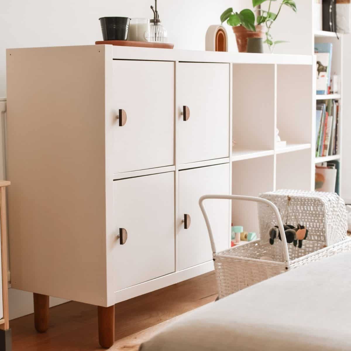 A white cabinet with wooden legs, resembling an IKEA Kallax hack, stands in a bright room. It features four closed compartments with circular handles and open shelves displaying small items. Nearby sits a wicker basket, while plants and books adorn the top and sides of the cabinet.