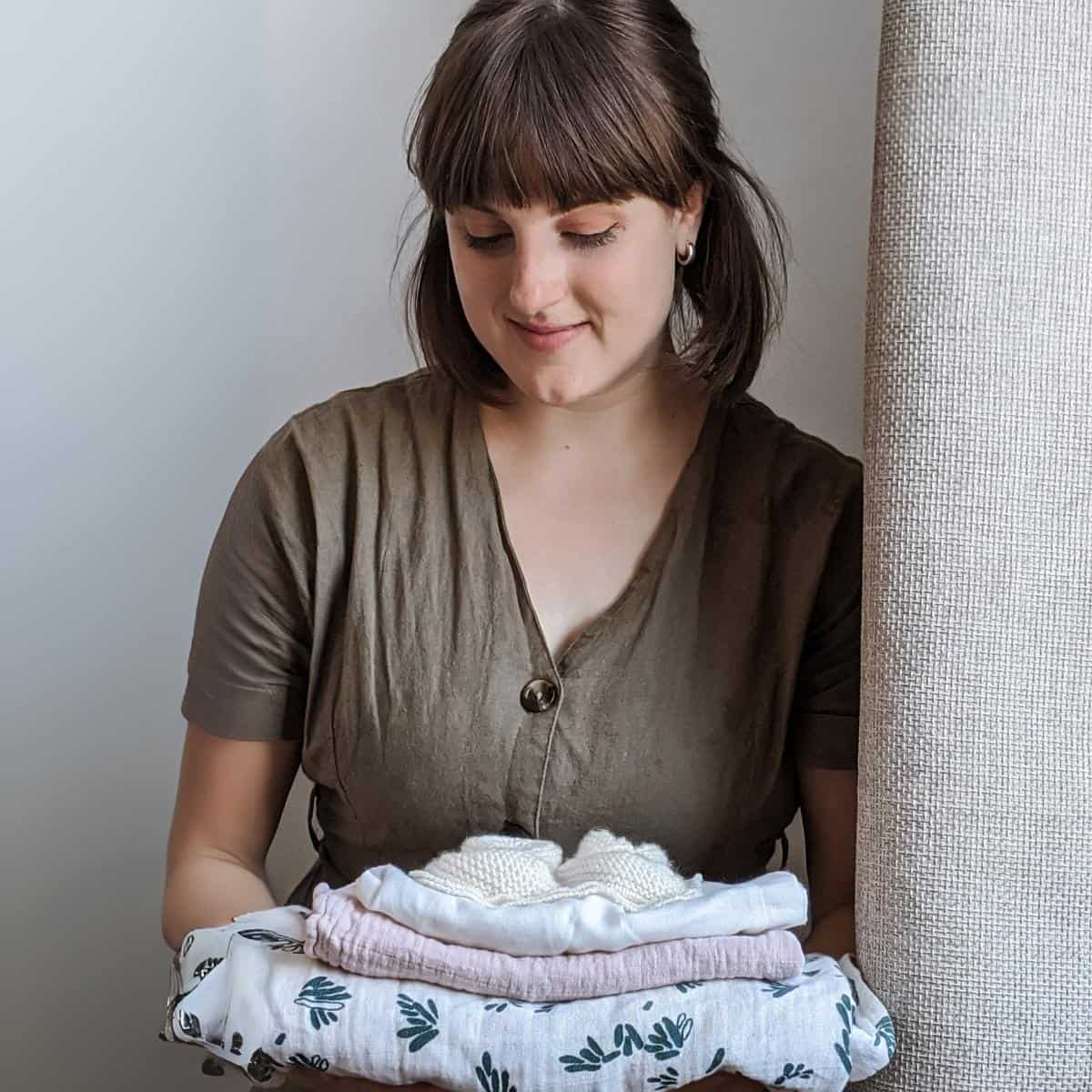 A woman with shoulder-length hair is holding a neatly folded stack of sustainable baby clothes, including a small pair of knitted shoes, while standing indoors near a curtain. Shes wearing a brown buttoned top and looking down at the clothes with care.