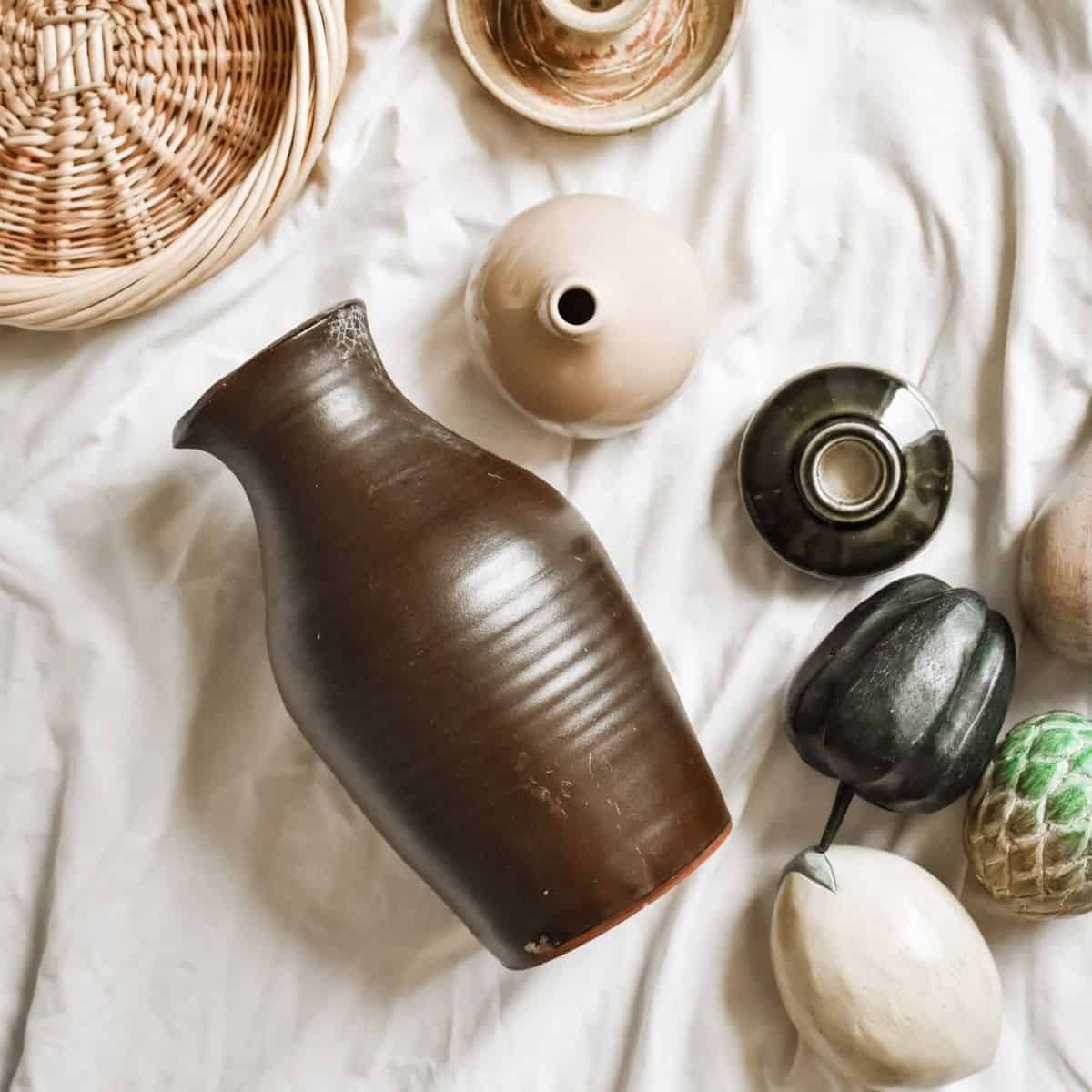 A curated collection of secondhand pottery and decorative items rests on a white fabric. A large brown vase takes center stage, surrounded by smaller vases and textured objects in neutral tones. A wicker basket peeks out at the top left, all in search of new places to call home.