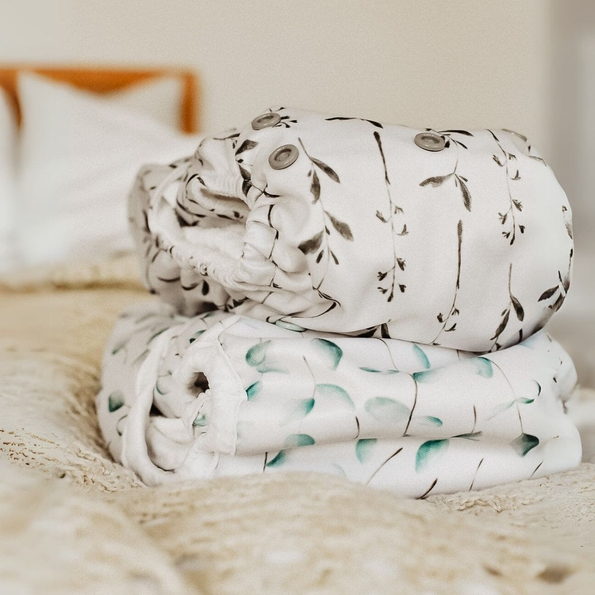 Two cloth diapers are stacked on a bed with a beige blanket. The top diaper, showcased in the review of Fiyyah, features a black and white leafy pattern, while the bottom one has a green leaf design. Pillows and a headboard are blurred in the background.