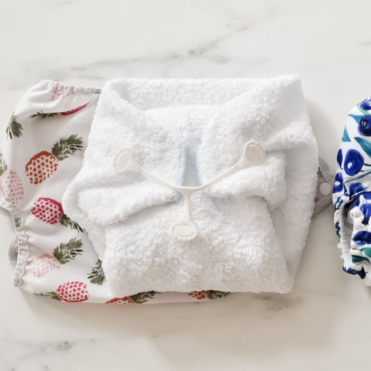 Cloth diapers are displayed on a white surface. The left one features a colorful fruit pattern, the middle is a fluffy white nighttime reusable nappy with a fastening clip, and the right has a blue and black pattern.