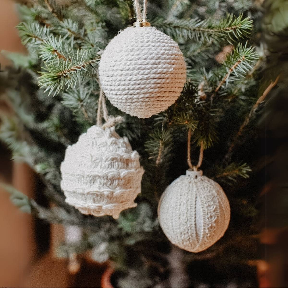 Three white, knitted ornaments, crafted from upcycled Christmas decorations, hang on a lush green tree. Each ornament boasts a unique texture: one with a cable knit pattern, another with a basket weave, and the third showcasing a ribbed design.