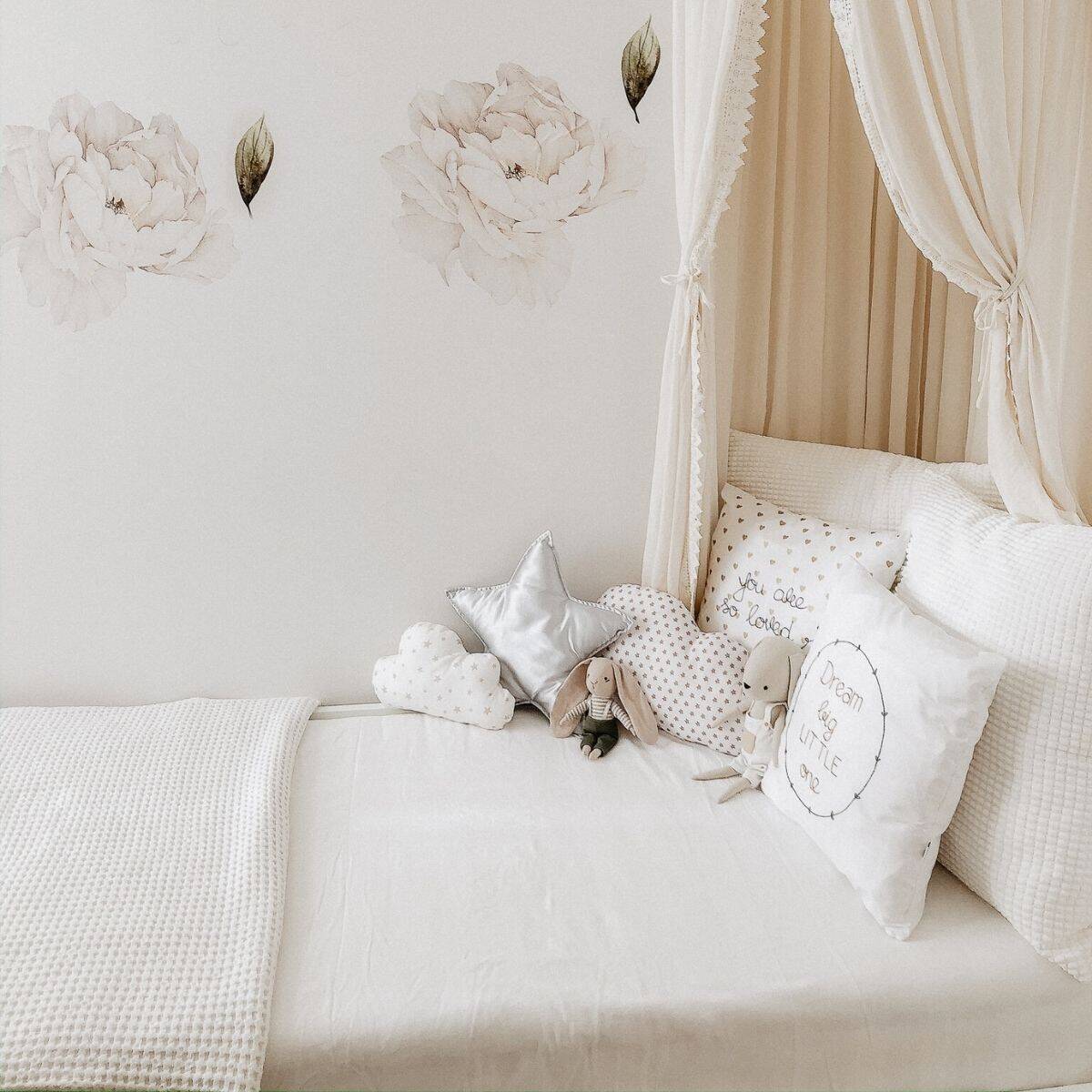 A cozy, light-themed bed with white netting canopy graces the sustainable bedroom. The bedspread is white with plush pillows, including star and cloud shapes. Floral wall decals adorn the wall, enhancing the serene ambiance of this eco-friendly space.