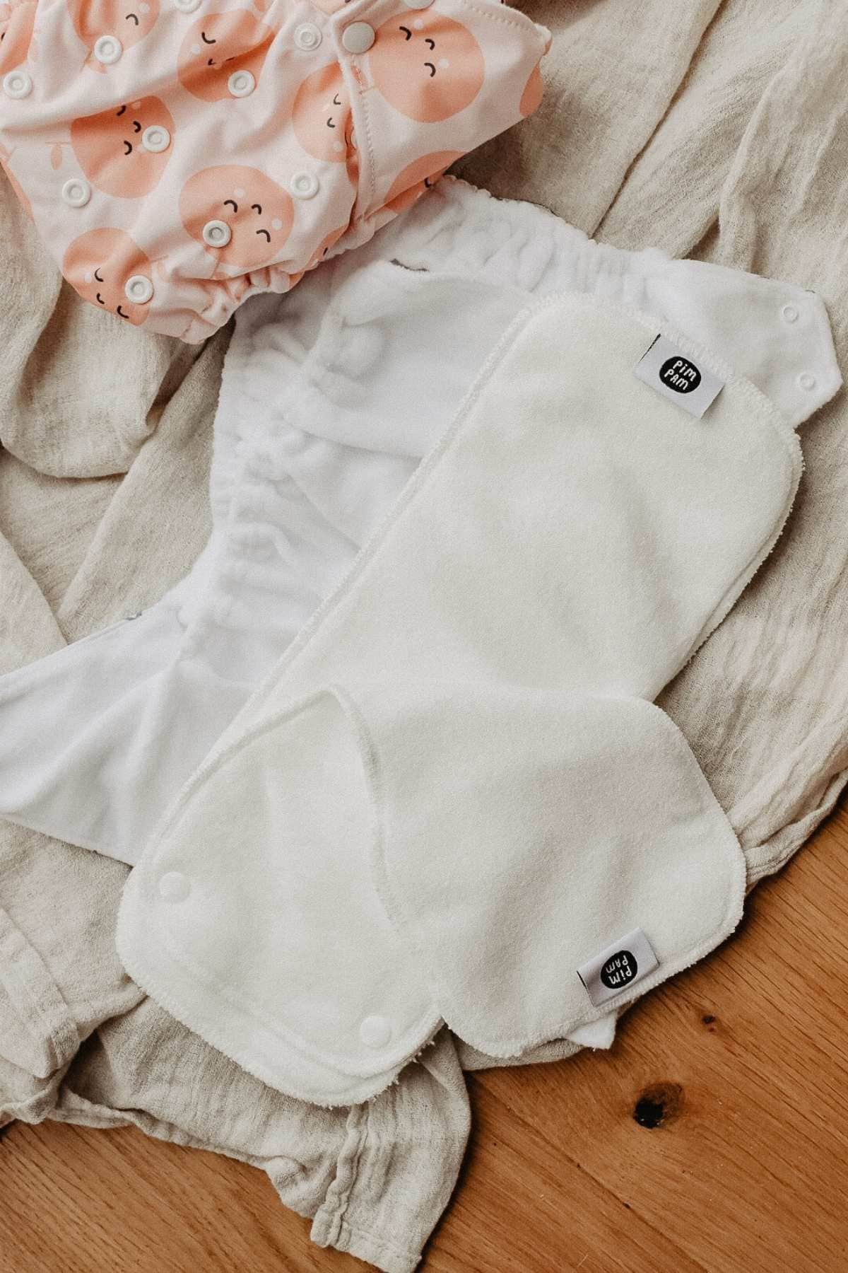 Cloth diapers and a reusable insert are laid out on a wooden surface. A pastel peach pocket nappy with happy faces stands out, showcasing an eco-friendly diapering option. Discover the convenience and cheerfulness of the Pim Pam Pocket Nappy in this charming review.