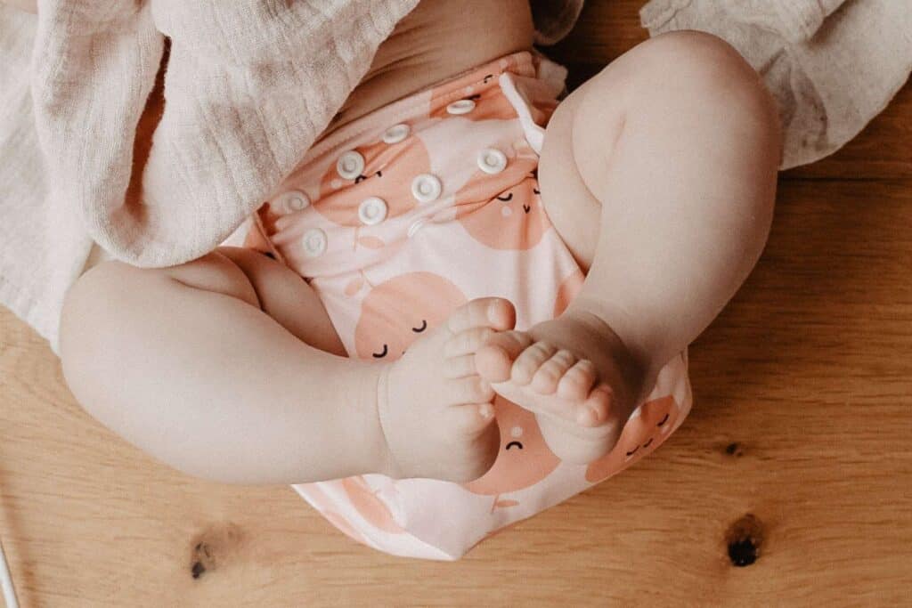 Close-up of a babys legs and feet, wearing a colorful cloth Pim Pam Pocket Nappy with a playful fruit pattern. The baby lies on a wooden surface, partially covered by a light fabric.