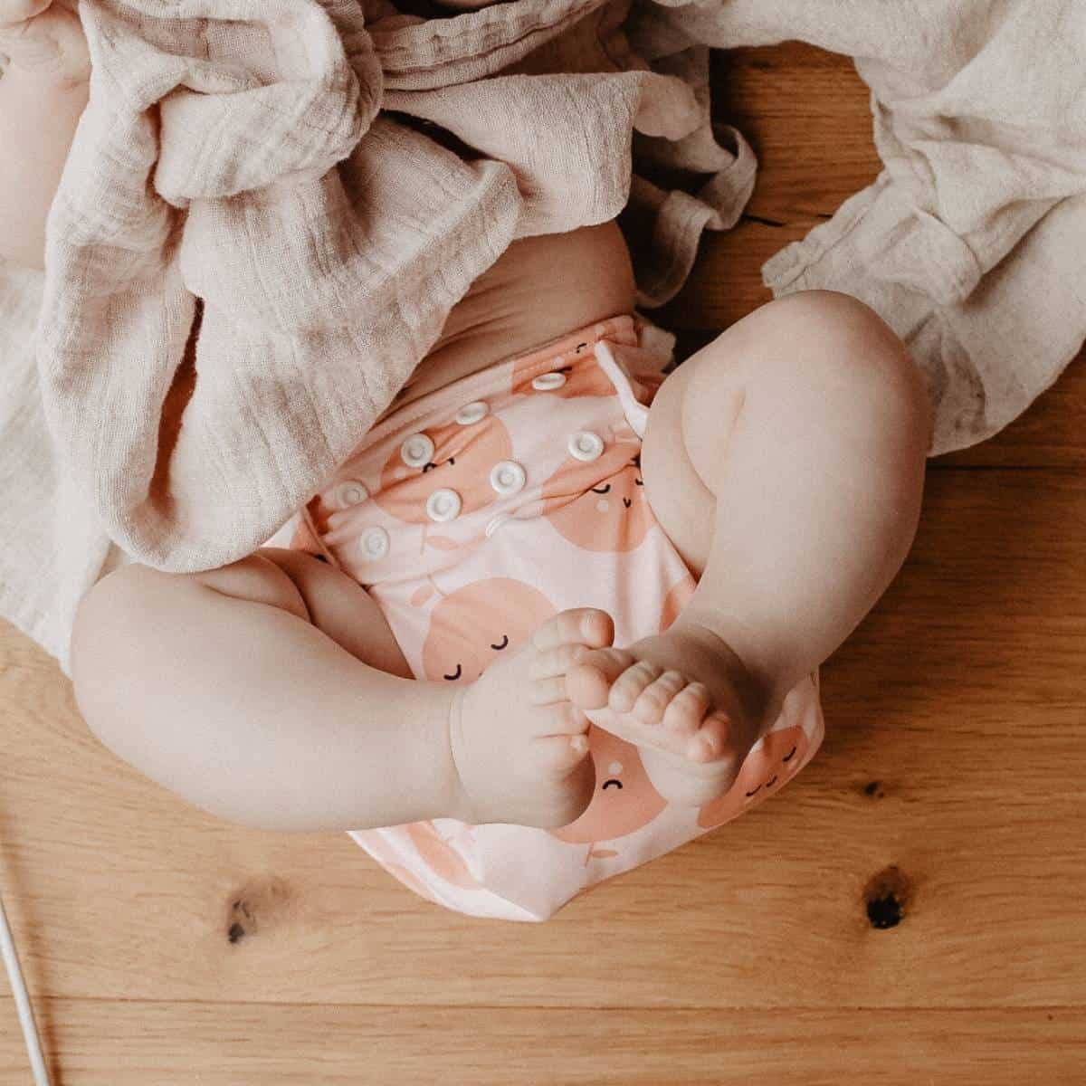 A baby lies on a wooden floor, wearing a Pim Pam Pocket nappy adorned with vibrant peach designs. Their legs are bent, and a light beige fabric is partially draped over them, showcasing the nappy's cheerful patterns.