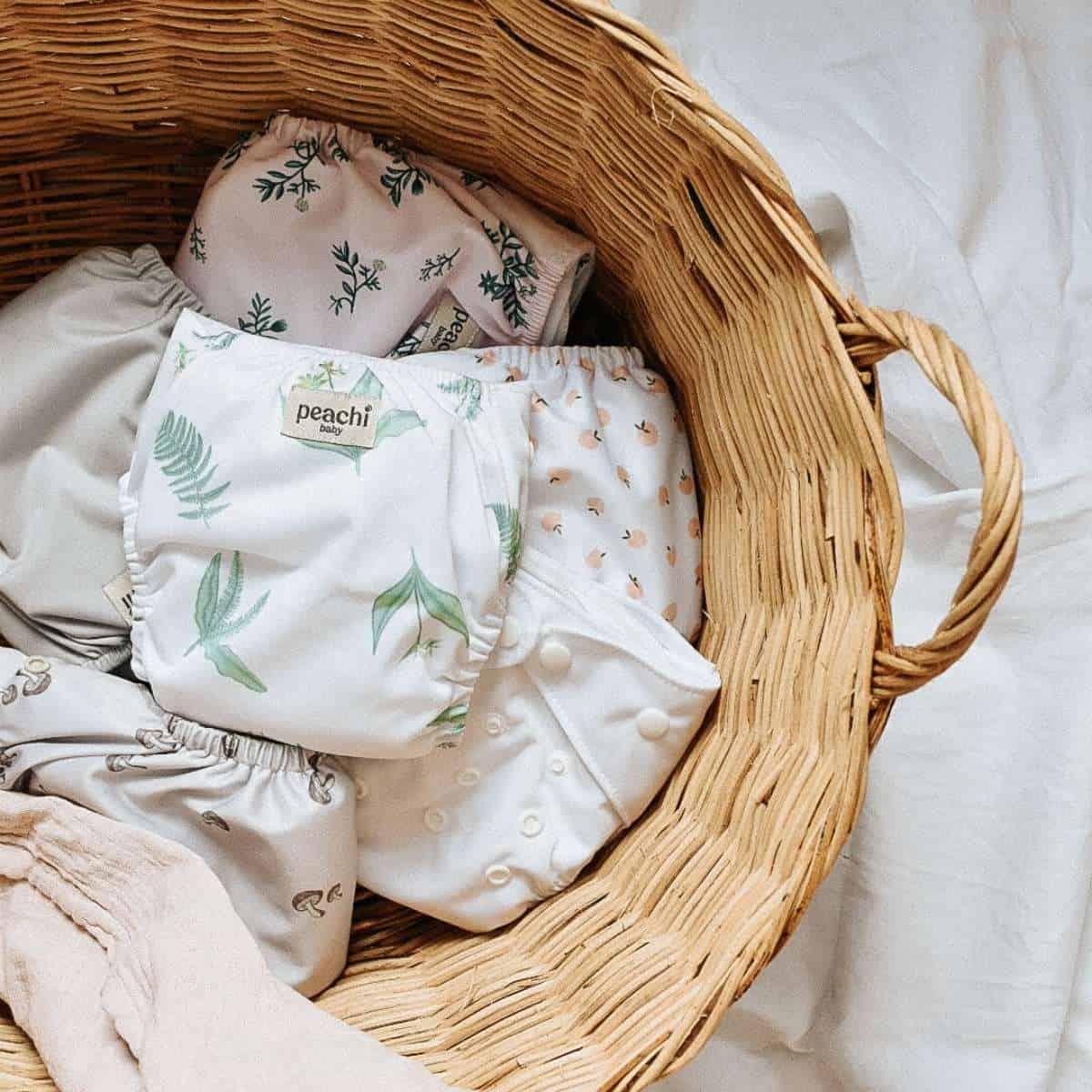 A wicker basket holds eco-friendly cloth diapers adorned with plant and floral patterns. The soft hues of white and cream blend seamlessly with green leafy designs. Perfect for conscious parents, this charming ensemble rests on a light-colored fabric surface, embracing sustainable choices for children.