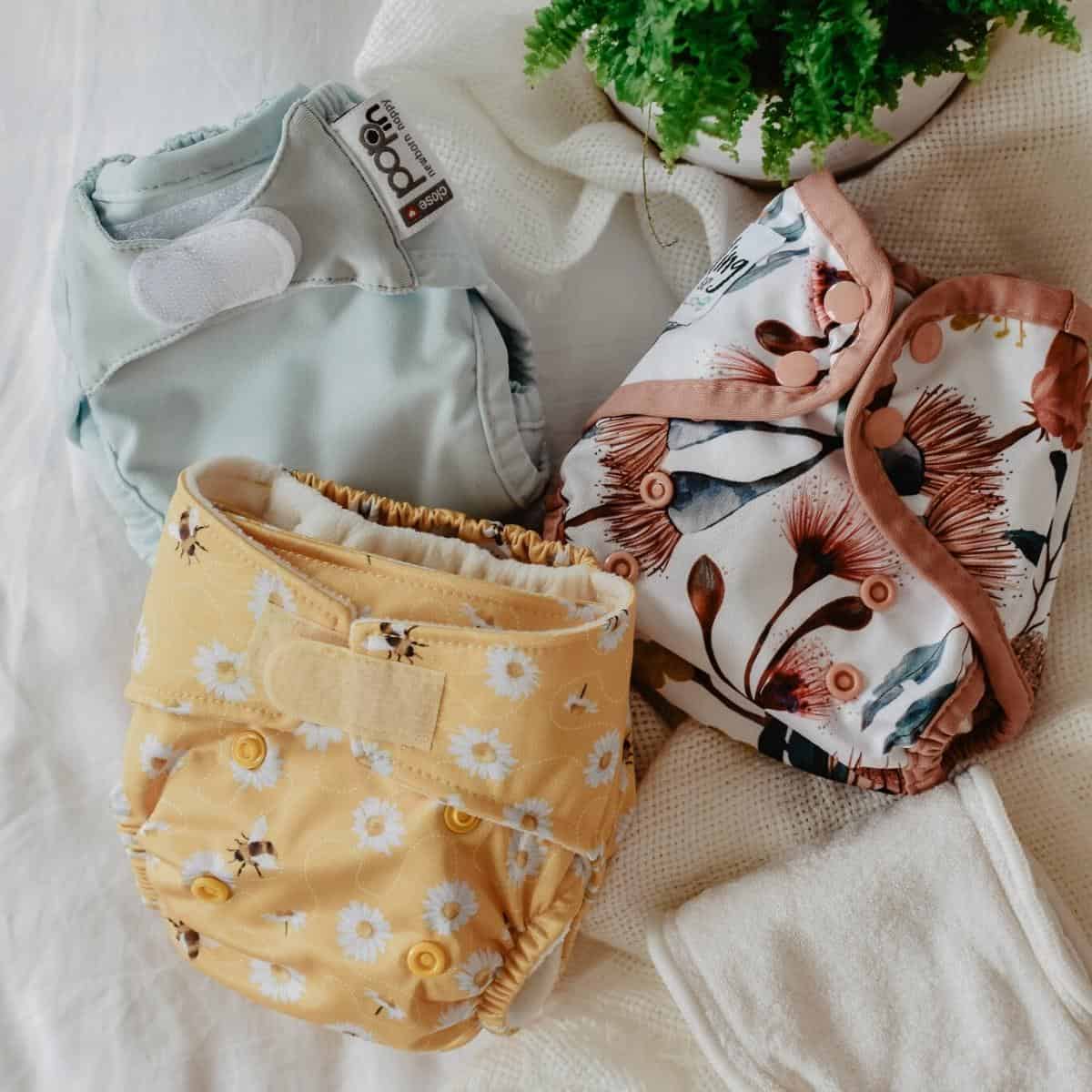 Three newborn reusable nappies in pastel blue, yellow with daisies, and a floral pattern are displayed on a white fabric surface. A green plant is visible at the top, adding a touch of nature.