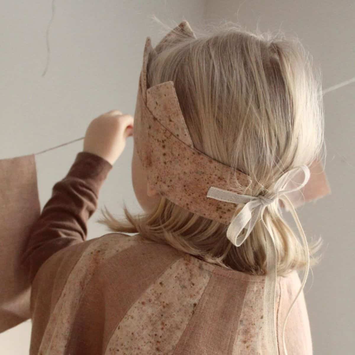 A child with long blonde hair, seen from behind, wearing a brown top and a textured, fabric crown. They reach up toward a string of eco-friendly fabric flags. The softly lit scene creates a warm, neutral atmosphere, perfect for thoughtful childrens moments.