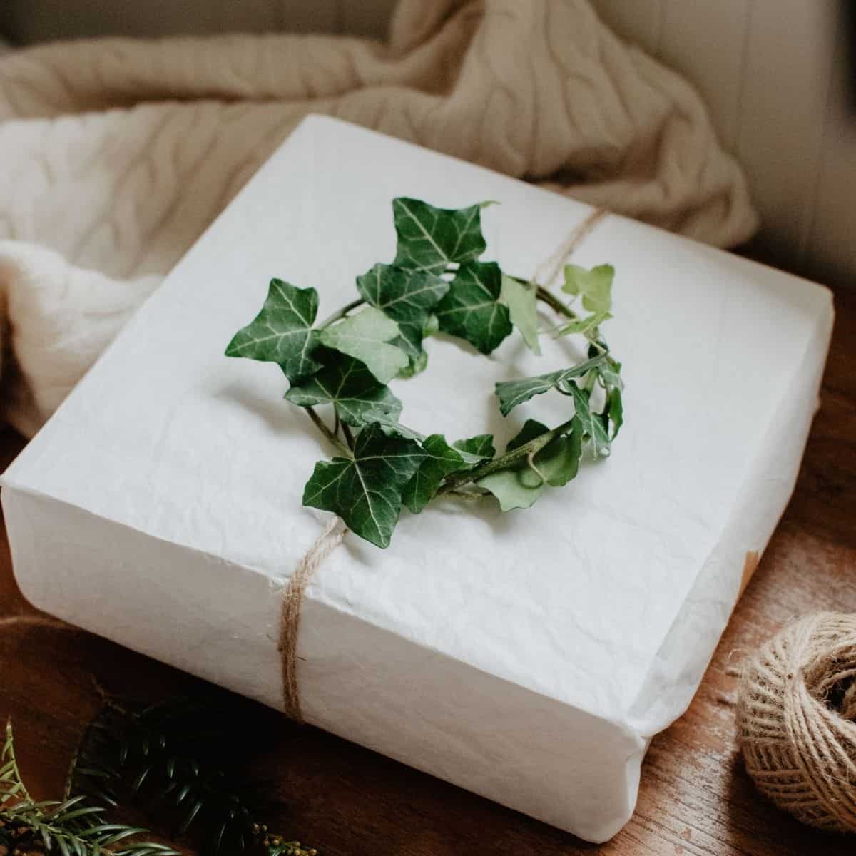 A gift wrapped in white paper is tied with twine, showcasing the charm of eco-friendly gifts. A small wreath of green ivy leaves adorns the package. Nearby, a spool of twine and some greenery rest on a wooden surface, complemented by a cream-colored cloth in the background.