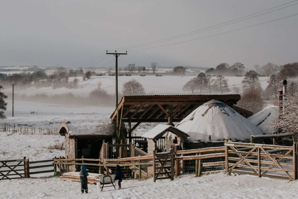 A snowy rural landscape features the Fordhall Farm glamping Yurts, surrounded by fences and fields. Two toddlers stand near a sled in the foreground. Rolling hills and bare trees are visible in the misty background.