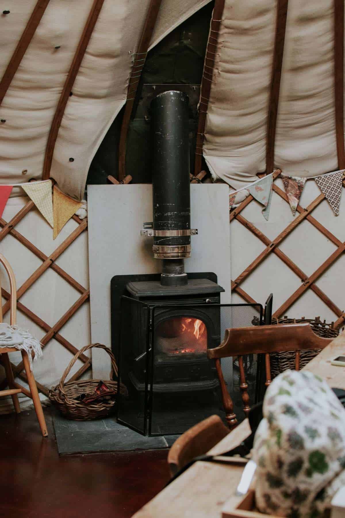 The cozy interior of a Fordhall Farm Yurt boasts a wood-burning stove with visible flames, keeping the yurt warm for small children and toddlers. Ceiling and walls are draped in fabric with wooden supports, while colorful bunting adds cheer. A rustic wooden chair and table sit prominently.