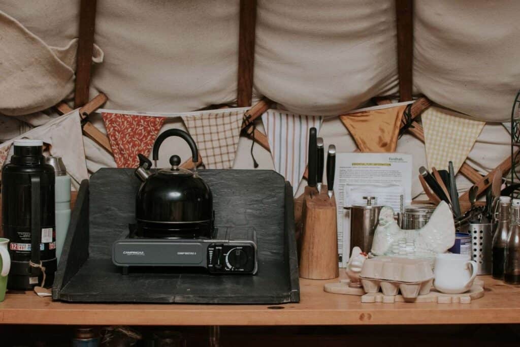 A cozy kitchen at Fordhall Farm Yurts features a black kettle on a portable stove, assorted kitchen utensils, and a wood block with knives. A black thermos and cloth bunting adorn the wooden counter.