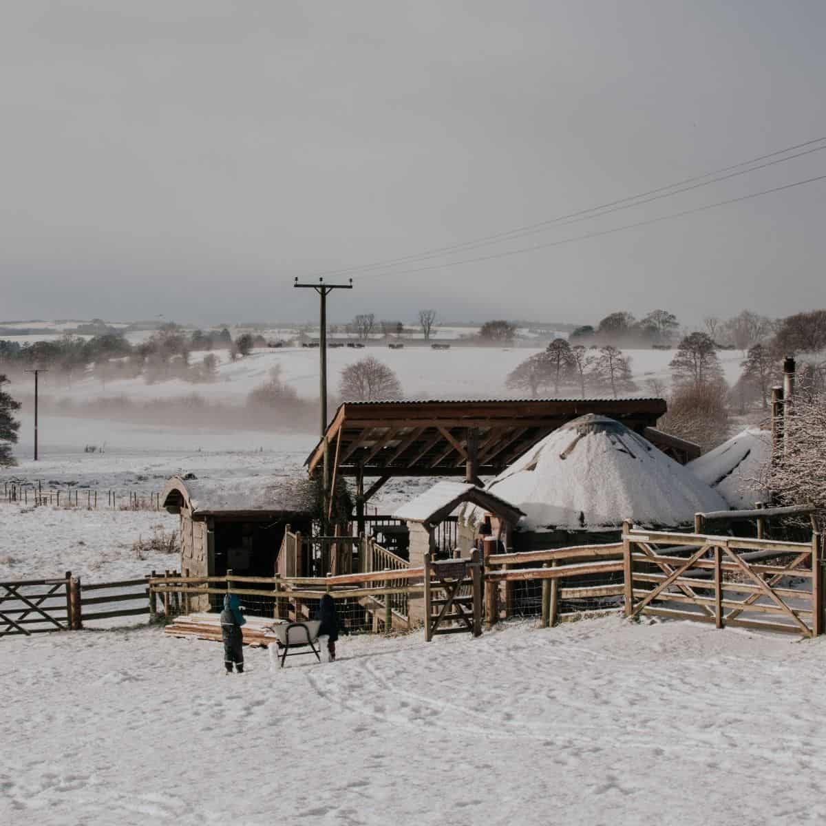 A snow-covered rural scene features the Fordhall Farm yurts. Two toddlers in winter clothing, walk through the snow toward the yurts. Leafless trees and rolling hills fade into the misty background under an overcast sky.