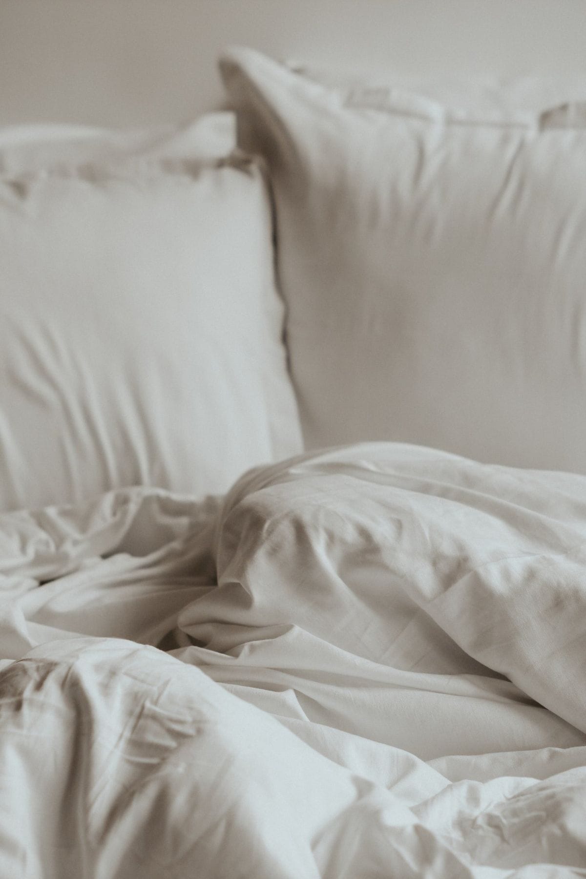 A close-up of an unmade bed with white pillows and crumpled sustainable bedding. The soft fabric appears inviting and cozy, conveying a sense of relaxation. The neutral tones create a calm, serene atmosphere.