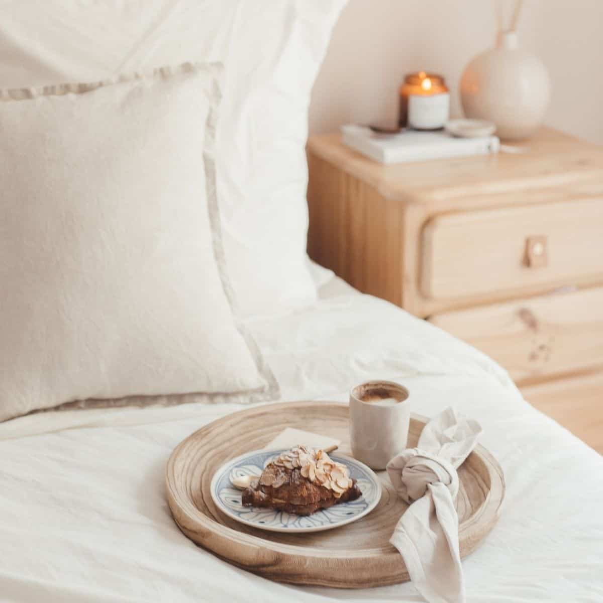 A cozy bedroom scene with a wooden tray on a white bed with sustainable bedding. The tray holds a pastry topped with almonds, a cup of coffee, and a beige napkin. A wooden nightstand with books, a vase, and a lit candle is in the background.