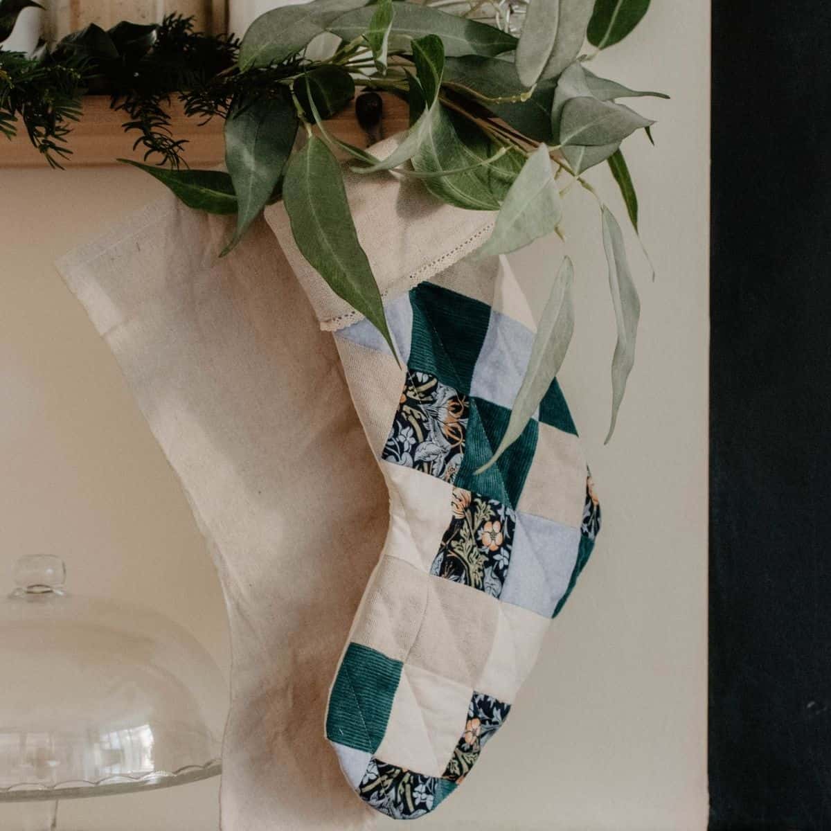 A patchwork stocking with green, blue, and patterned squares hangs beside a plain beige stocking. Both are adorned with green foliage at the top and dangle on a wall near a glass dome.
