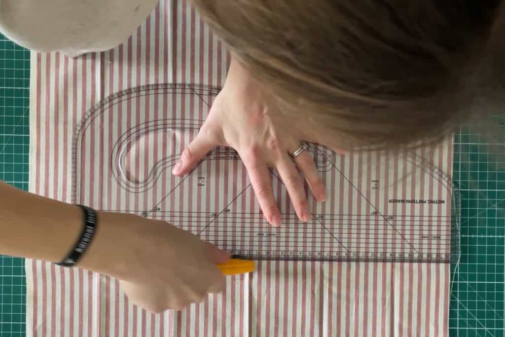 Person using a rotary cutter and transparent ruler on red and white striped fabric on a green cutting mat. Fabric is neatly aligned with grid, indicating precision in cutting.