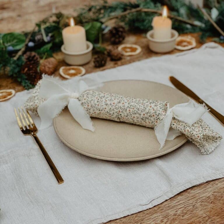 A floral reusable christmas cracker lying on a plate with greenery and candles in the background