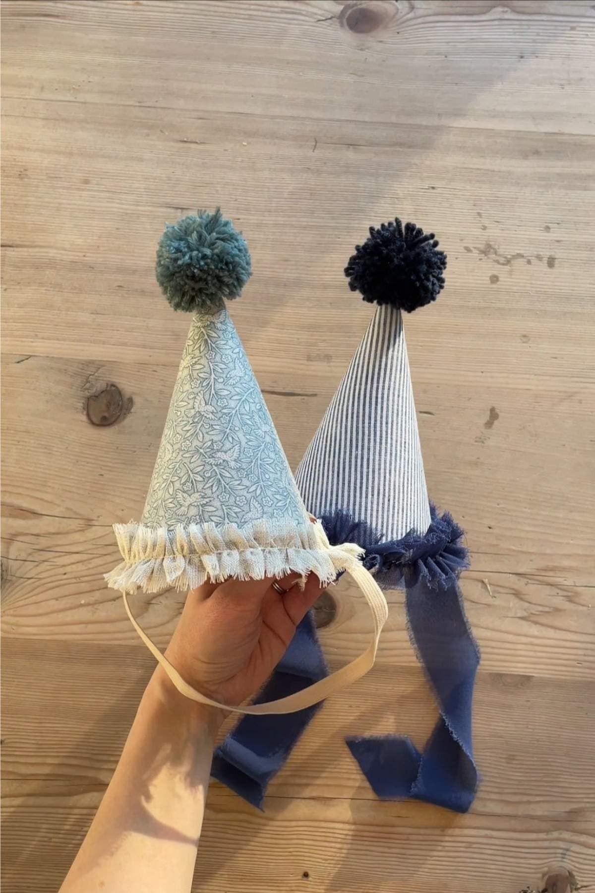 A hand holds two whimsical DIY party hats with ribbons. One is light blue with a green pom-pom, while the other has blue and white stripes with a navy pom-pom. 