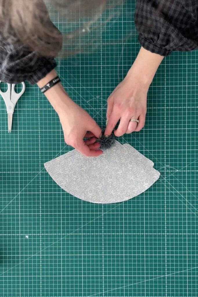 A person is crafting DIY party hats on a green cutting mat, applying the pom pom to the top of the hat. Scissors lay nearby.