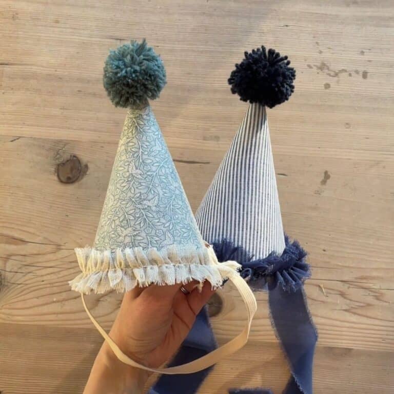 Two handmade party hats are held up against a wooden surface. Each cone-shaped hat features a pom-pom on top, ruffled trim at the base, and ribbon ties.