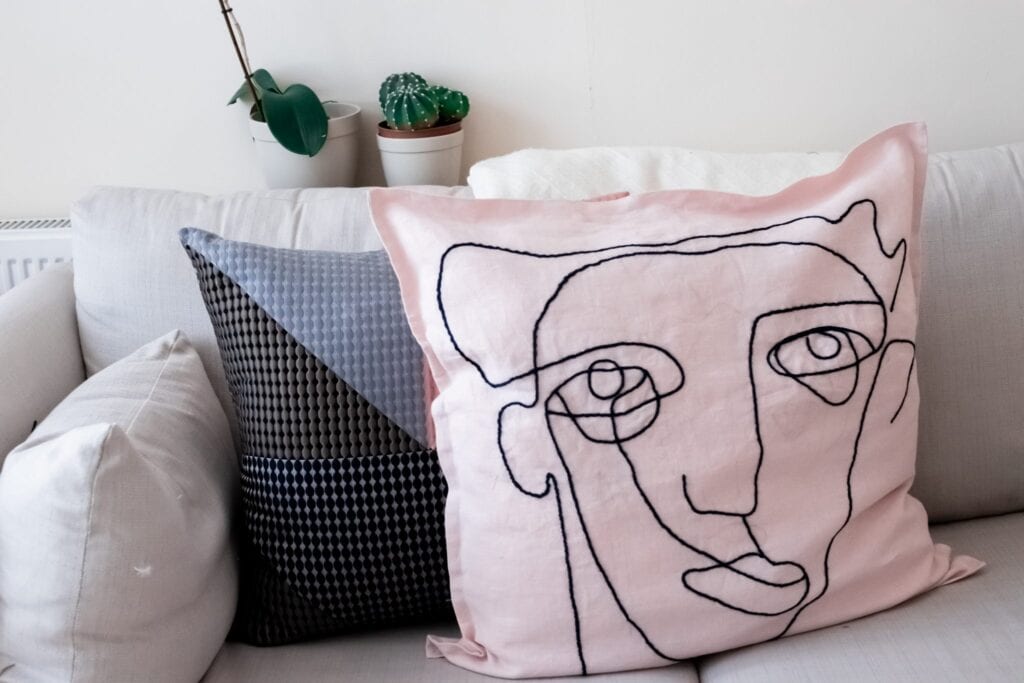A cozy couch features three decorative cushions, including an embroidered pink one with a black abstract face design and another with a black and white geometric pattern. A simple white abstract cushion complements the set, while two small potted plants make their home on the shelf above.