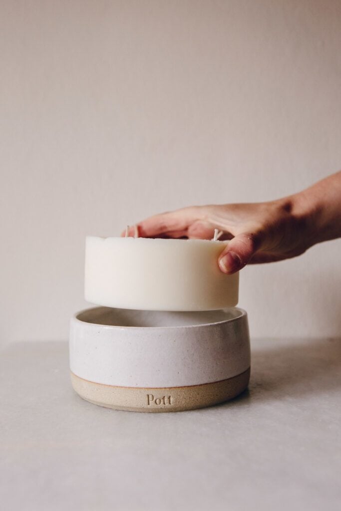 A hand lifts a white, round wax block with wicks from a ceramic bowl labeled Pott. The bowls matte texture complements the light surface and plain background, making it an ideal eco-friendly gift for her or him.