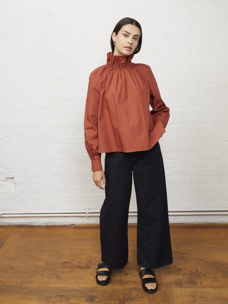 A person stands against a white brick wall in a rust-colored, high-neck blouse and wide-leg black pants. With relaxed hands by their sides and black sandals on their feet. Their long dark hair frames a serene expression.
