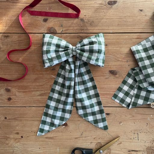 A handmade gingham fabric bow lies on a rustic farmhouse table with red ribbon and fabric shears