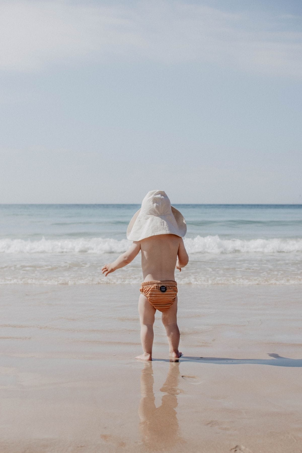 A toddler wearing a sun hat and striped swim shorts, ready for a holiday adventure, stands at the edge of the beach. The clear sky and gentle waves kiss the sandy shore as he enjoys his day, with a reassuringly eco-friendly cloth nappy beneath his trendy outfit.