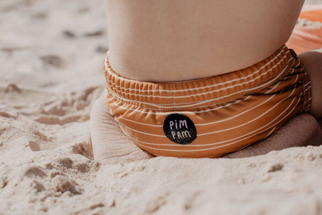 A child sitting on a sandy beach during a holiday, wearing orange, striped swim trunks with PIM PAM on a circular patch.
