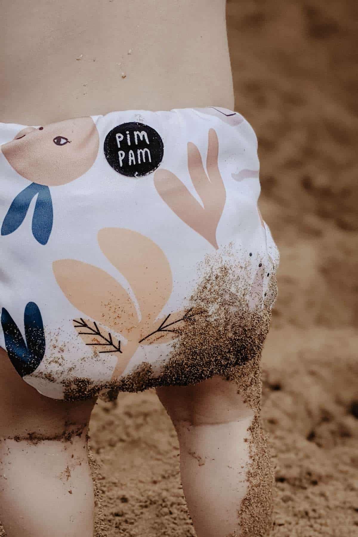 A baby with sandy legs stands on the beach, wearing a white cloth nappy adorned with abstract designs of shapes and faces, perfect for a holiday outing. The patch on the nappy reads Pim Pam. The background is a soft sandy surface.