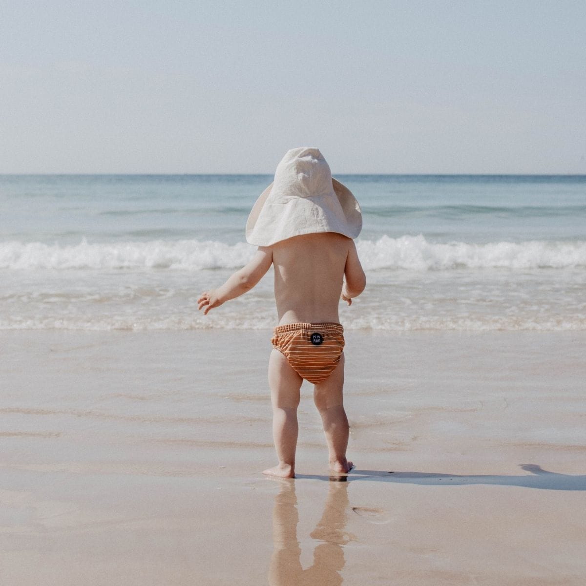 A toddler in a wide-brimmed hat and orange-striped cloth nappy stands at the oceans edge, facing the waves on a sunny holiday beach day. The clear sky stretches above as the water gently laps at their feet.