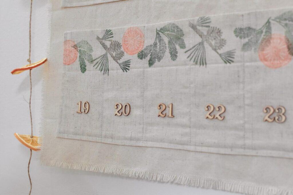 Close-up of a DIY fabric calendar featuring dates 19 to 23 with hand-stamped illustrated greenery and orange slices. This advent calendar is hung on a wall, with dried orange slices at the bottom left corner.