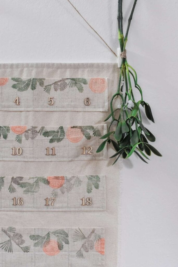 A fabric Advent calendar with numbered pockets, decorated with hand-stamped green and orange botanical patterns. It hangs from a string, featuring a bundle of green leaves attached at the top right corner.