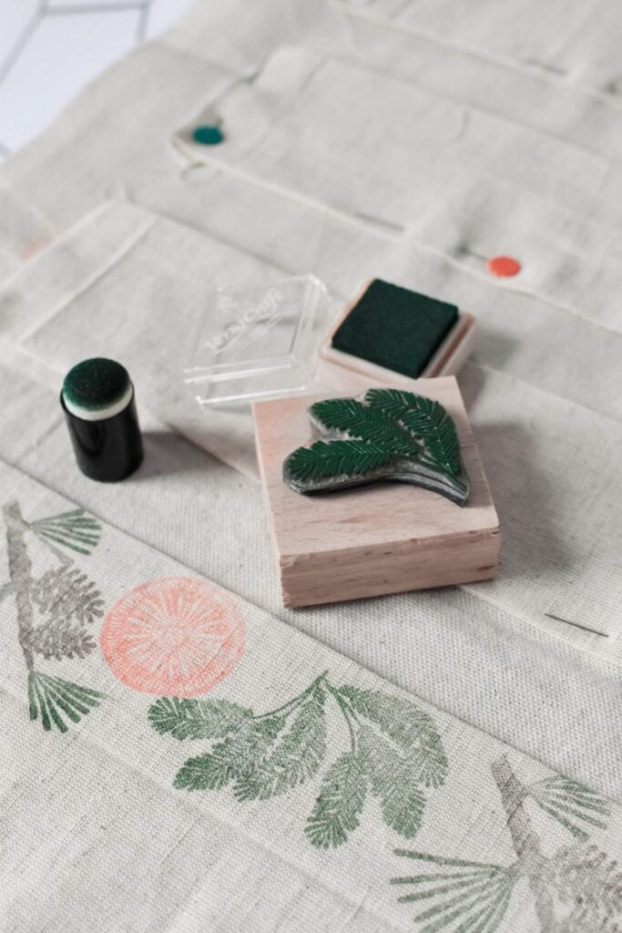 A fabric with leaf and orange stamped patterns is displayed. Nearby, wooden stamps for hand-stamped designs feature leaf imprints and ink pads in orange and green, perfect for a DIY advent calendar project. A green stamp press lies on a light-colored surface.