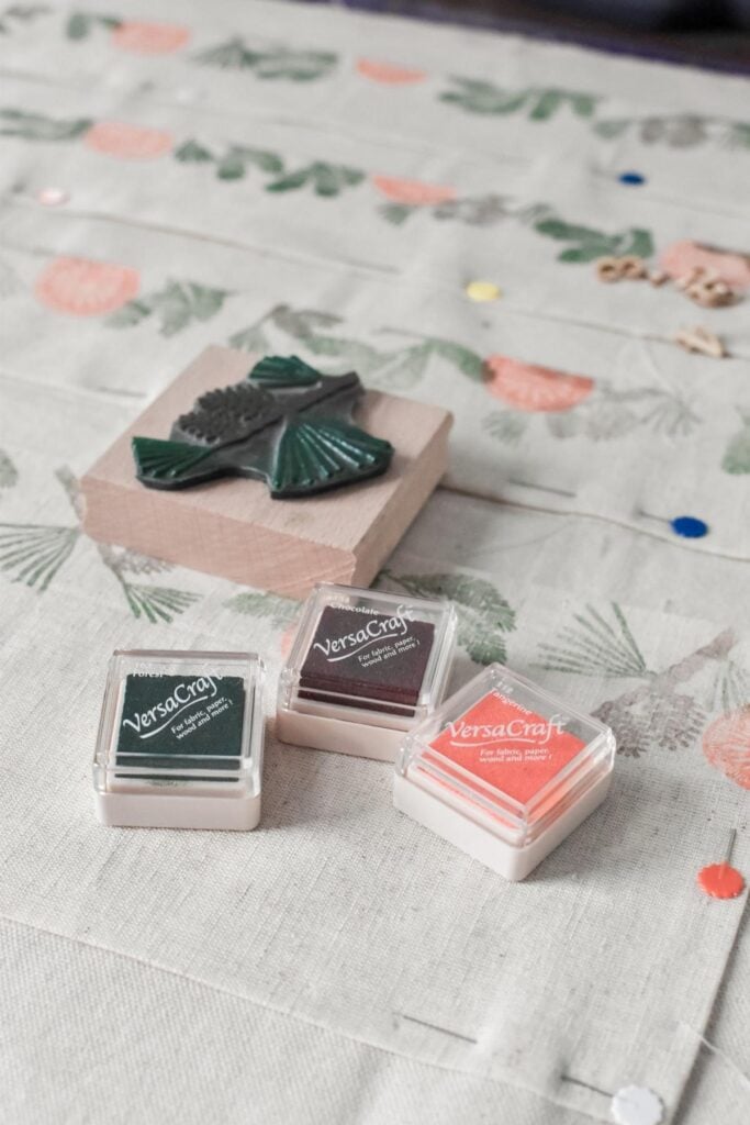 Crafting materials are displayed on a patterned handmade advent calendar. Three VersaCraft ink pads in green, dark gray, and coral sit beside a stamp with a wooden leaf design. 