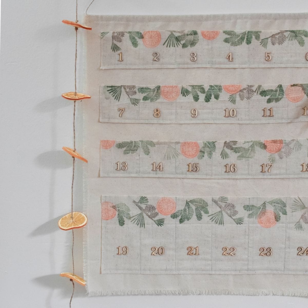 This hand-stamped fabric wall calendar with floral and leaf patterns features days 1 to 31 in brown. Hung on twine, it is adorned with dried orange slices attached by small clothes pins.