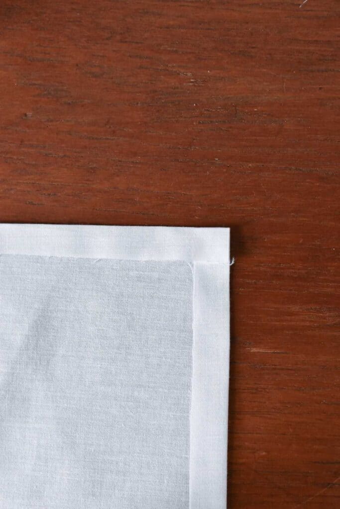 Close-up of a white fabric placed on a wooden surface, perfect for creating beautiful handkerchiefs. The fabric is neatly folded at the corner, showcasing a clean edge against the rich brown texture of the wood.