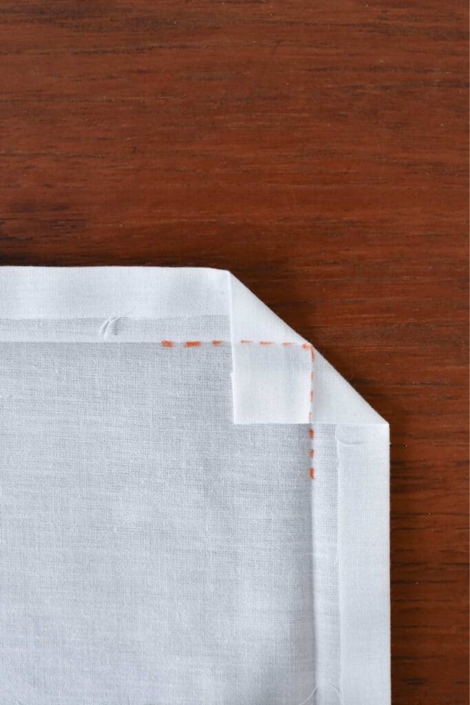 Close-up of a neatly folded white fabric with visible orange stitching, placed on a wooden surface. The beautiful handkerchiefs corner is carefully aligned, showcasing precise craftsmanship.
