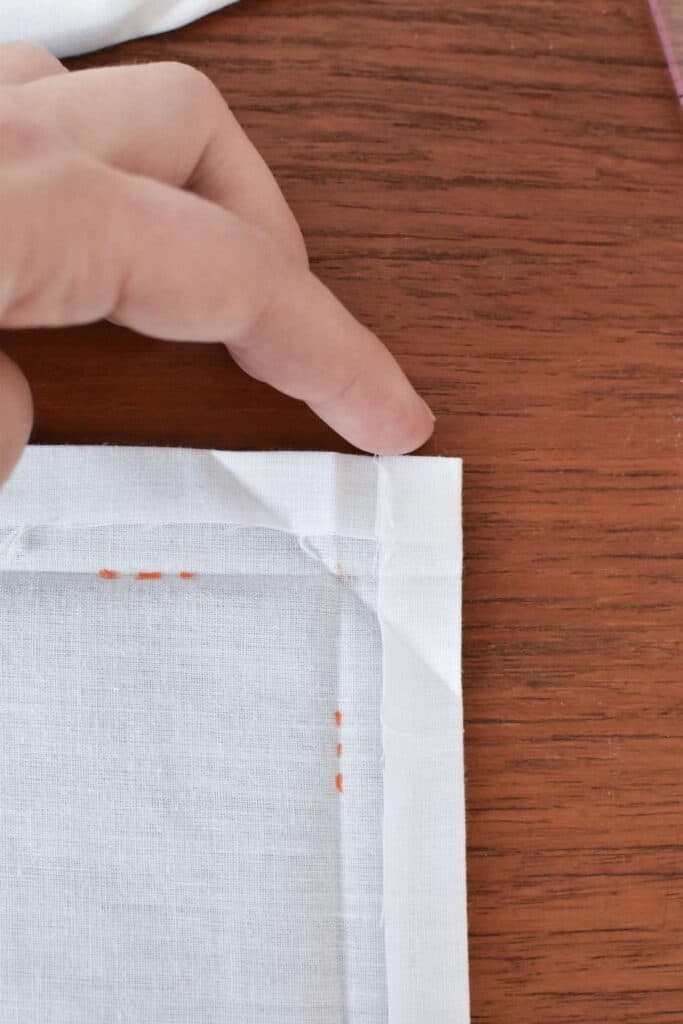 Close-up of a person’s hand pressing the corner of a white fabric, laid out on a wooden surface, showcasing visible stitching along the edges. The fabric looks like its in the process of becoming beautiful handkerchiefs, crafted with care.