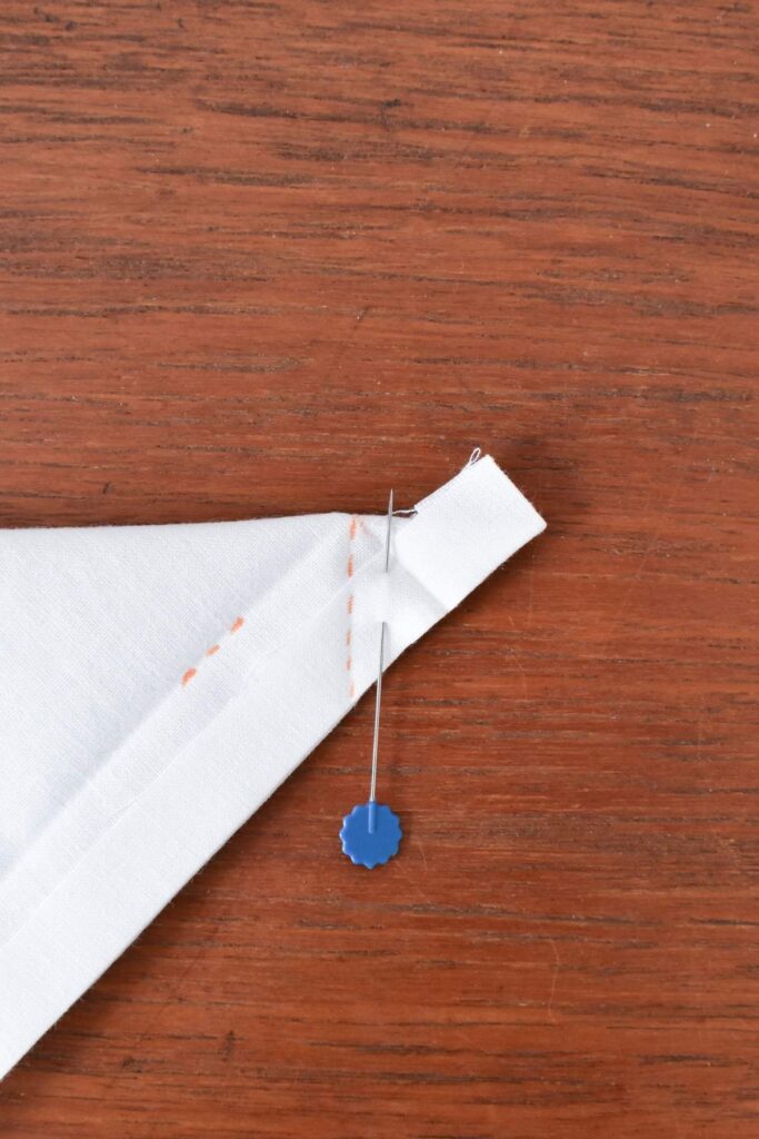 A beautiful white handkerchief, folded into a triangular shape, is pinned with a straight pin featuring a blue head. It boasts orange stitching along one edge and rests effortlessly on a wooden surface.
