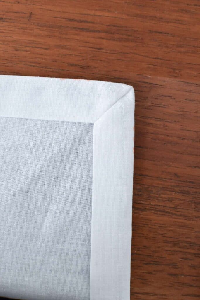 Close-up of a beautifully folded corner of a light blue fabric napkin on a wooden table. The smooth, clean fabric creates a sharp, tidy appearance against the warm brown wood grain background.